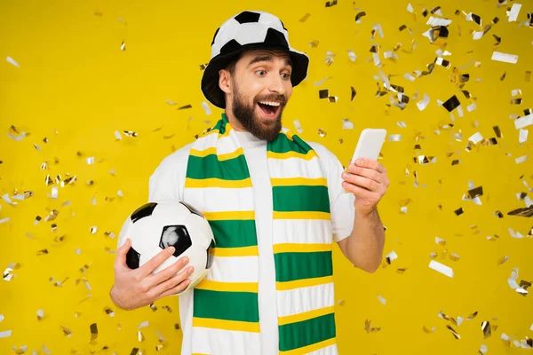 Admirador de los deportes sorprendidos y felices con pelota de fútbol viendo el partido de fútbol en el teléfono inteligente cerca de confeti en amarillo - foto de stock