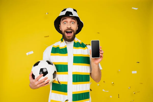 Изумленный спортивный болельщик в полосатом шарфе с футбольным мячом и смартфоном с чистым экраном на желтом — стоковое фото