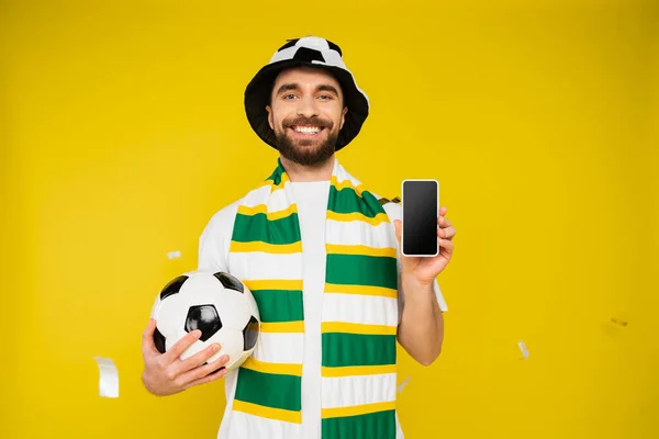 Fröhlicher Sportfan mit Fußballball zeigt Smartphone mit leerem Bildschirm auf gelbem Hintergrund — Stockfoto