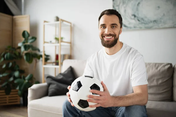 Bärtiger Sportfan sitzt auf Couch im verschwommenen Wohnzimmer und lächelt in die Kamera — Stockfoto