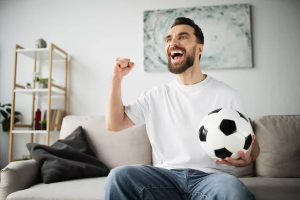 Emocionado fanático del fútbol sosteniendo la pelota y mostrando el gesto de triunfo mientras ve el partido en la sala de estar en casa - foto de stock