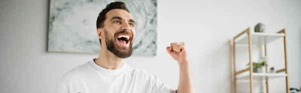Задоволений спортивний фанат кричить і показує жест виграшу під час перегляду гри вдома, банер — стокове фото