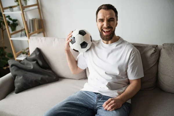 Схвильований футбольний фанат сидить на дивані з футбольним м'ячем і дивиться на камеру — стокове фото