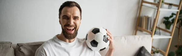 Возбужденный спортивный болельщик держит футбольный мяч, глядя на камеру дома, баннер — стоковое фото