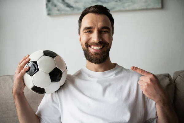 Happy bearded man looking at camera and pointing at soccer ball at home — Stock Photo