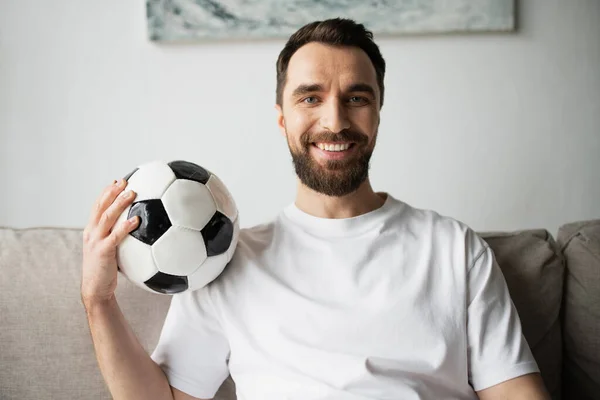 Весёлый и бородатый футбольный фанат держит футбольный мяч и улыбается в камеру — стоковое фото