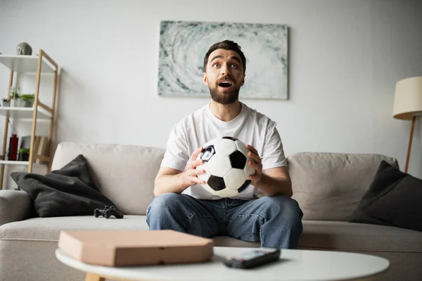 QUIIV, UCRÂNIA - OUTUBRO 21, 2022: homem maravilhado segurando futebol e assistindo campeonato em casa — Fotografia de Stock
