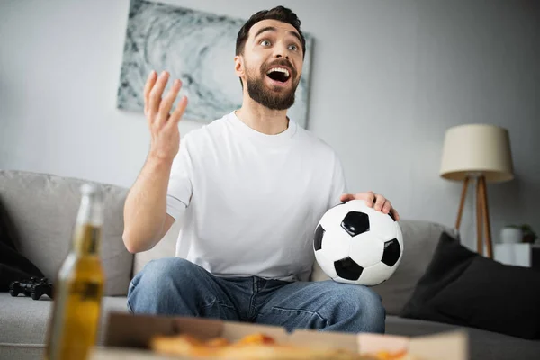 QUIIV, UCRÂNIA - OUTUBRO 21, 2022: homem alegre segurando futebol e gesticulando enquanto assiste ao campeonato em casa — Fotografia de Stock
