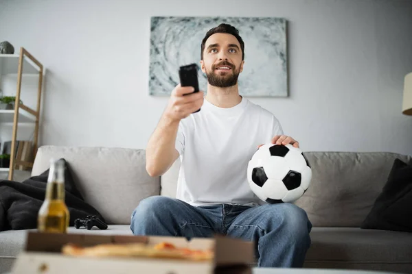 KYIV, UCRAINA - 21 OTTOBRE 2022: uomo allegro che tiene il calcio e il telecomando mentre guarda il campionato a casa — Foto stock
