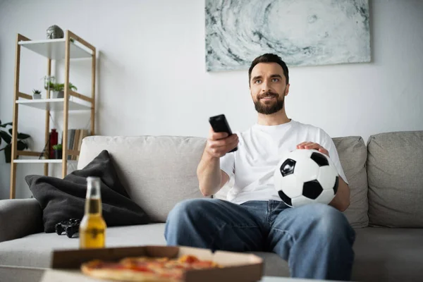 KYIV, UKRAINE - OCTOBER 21, 2022: happy man holding remote controller and football while watching championship at home — Stock Photo
