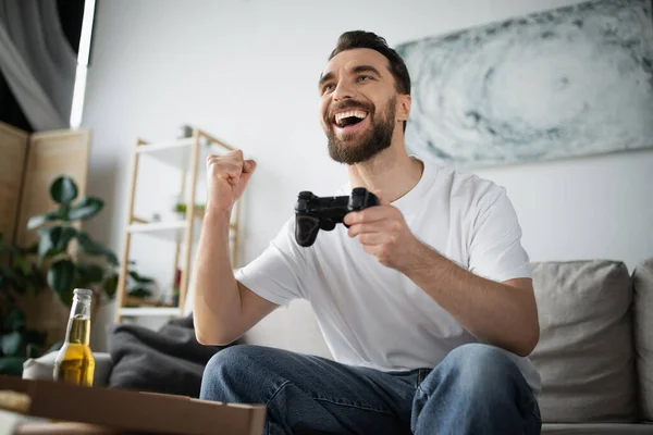 KYIV, UKRAINE - OCTOBER 21, 2022: excited man playing video game near pizza and bottle of beer at home — Stock Photo