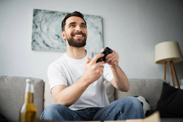 KYIV, UCRÂNIA - 21 de outubro de 2022: homem barbudo feliz jogando videogame perto de garrafa de cerveja em casa — Fotografia de Stock