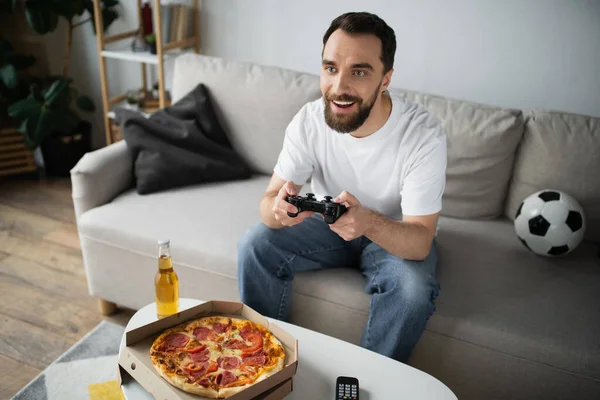 KYIV, UCRANIA - 21 de octubre de 2022: un hombre alegre jugando un videojuego cerca de una pizza y una botella de cerveza en casa - foto de stock