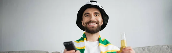 Hombre feliz en el sombrero del ventilador y la bufanda que sostiene el mando a distancia y la botella de cerveza mientras que mira el partido de fútbol, bandera - foto de stock