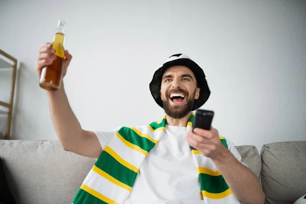 Alegre hombre en abanico sombrero y bufanda celebración mando a distancia y botella de cerveza mientras mira partido de fútbol - foto de stock