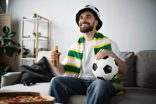 KYIV, UCRANIA - 21 DE OCTUBRE DE 2022: hombre alegre en sombrero y bufanda sosteniendo fútbol y botella de cerveza mientras ve el campeonato - foto de stock