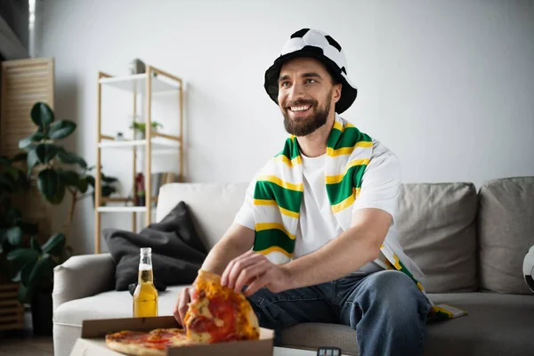 Веселый человек в шляпе и шарфе берет кусочек вкусной пиццы во время просмотра чемпионата — стоковое фото