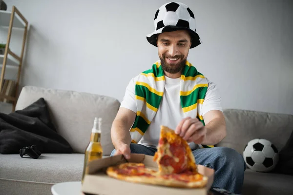 KYIV, UKRAINE - OCTOBER 21, 2022: joyful man in hat and scarf taking slice of tasty pizza during championship — Stock Photo