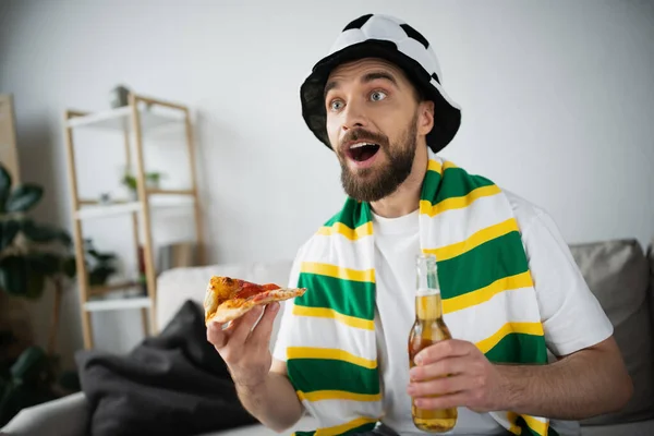 Erstaunter Mann mit Hut hält eine Scheibe leckere Pizza und eine Flasche Bier in der Hand, während er die Meisterschaft verfolgt — Stockfoto