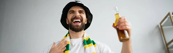 Вражений фанат спорту в шарфі і капелюсі тримає пляшку пива під час перегляду чемпіонату, банер — стокове фото