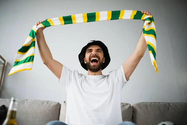 Збуджений чоловік у фан-капелюсі тримає натягнутий шарф під час крику і перегляду чемпіонату — стокове фото