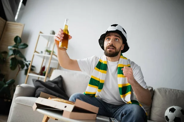 Tensed man in sportive fan hat and scarf holding bottle of beer while watching championship — Stock Photo