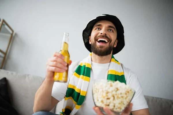 Положительный спортивный фанат в шляпе и шарфе, держащий миску с попкорном и бутылкой пива во время просмотра чемпионата — стоковое фото