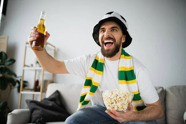 Возбужденный фанат спорта в шляпе и шарфе, держащий миску с попкорном и бутылкой пива во время просмотра чемпионата — стоковое фото