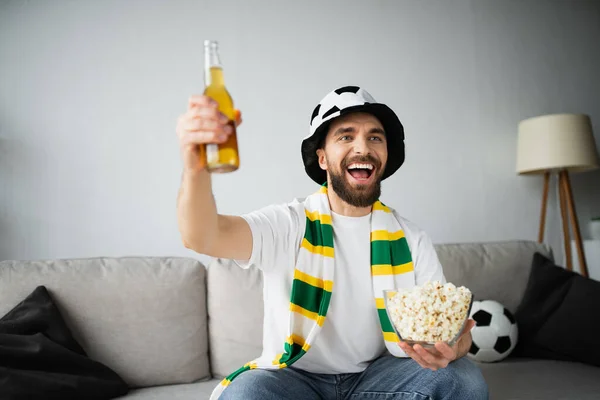 Fan de sport gai dans un chapeau et une écharpe tenant bol avec pop-corn et bouteille de bière tout en regardant le championnat — Photo de stock