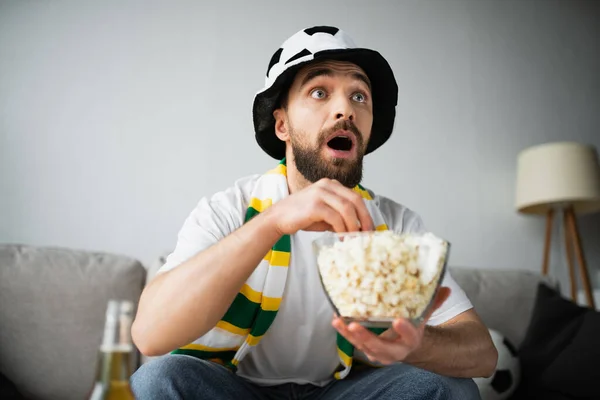 Sorprendido y barbudo hombre en deportivo abanico sombrero y bufanda celebración bowl con palomitas de maíz mientras mira campeonato - foto de stock