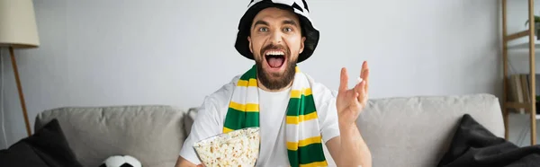 Homme excité en chapeau de ventilateur et foulard regarder championnat tout en tenant bol avec pop-corn, bannière — Photo de stock