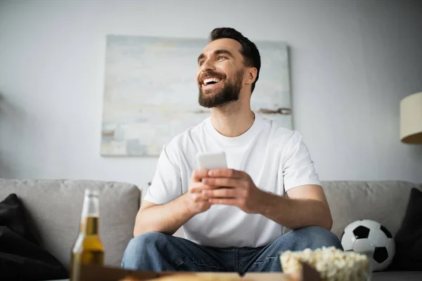Hombre alegre usando teléfono inteligente cerca de la botella de cerveza y comida sabrosa en primer plano borrosa - foto de stock