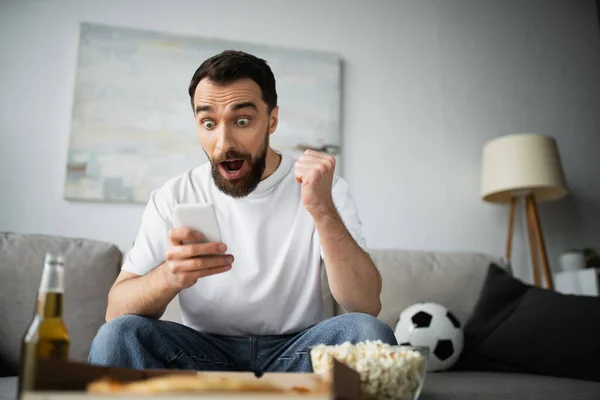 Hombre emocionado comprobar la puntuación de fútbol en el teléfono inteligente cerca de botella de cerveza y comida sabrosa en primer plano borrosa - foto de stock