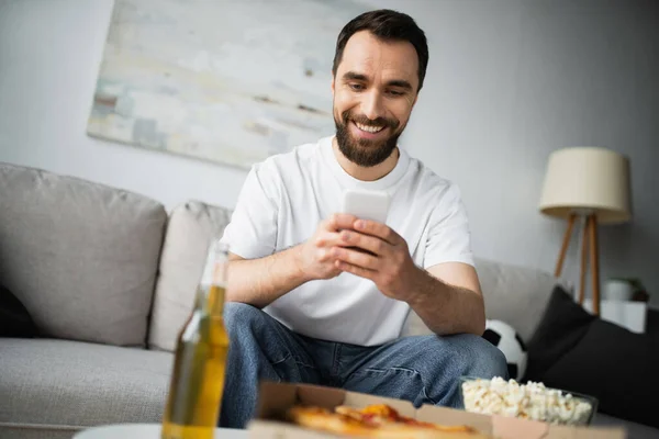 Sonriente hombre barbudo usando teléfono inteligente cerca de la botella de cerveza y comida sabrosa en primer plano borrosa - foto de stock