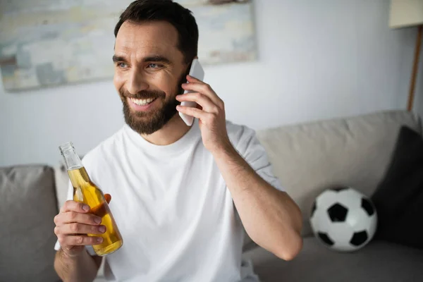 Веселый человек держит бутылку пива и разговаривает на смартфоне рядом с футболом на размытом диване — стоковое фото
