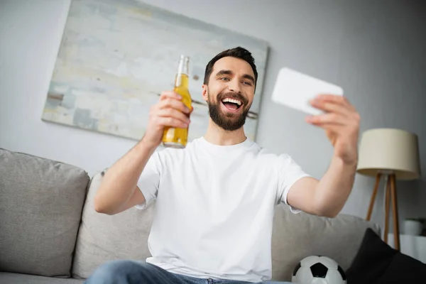 Веселый мужчина держит бутылку пива и делает селфи на смартфоне дома — Stock Photo