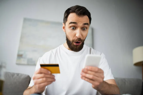 Sorprendido hombre que sostiene la tarjeta de crédito y el uso de teléfono inteligente mientras hace compras en línea en casa - foto de stock