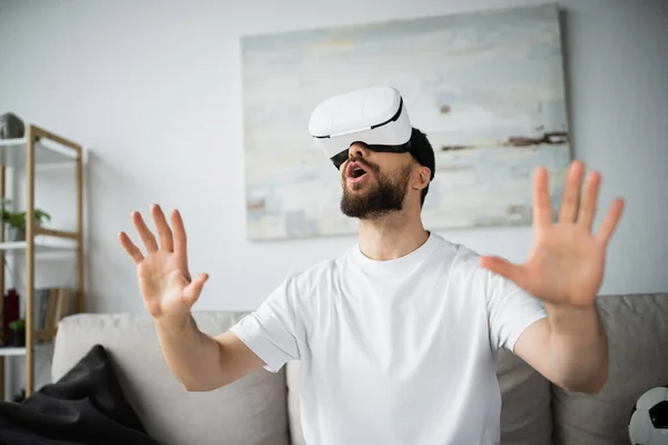 Ошеломленный и босый мужчина в гарнитуре VR жестикулирует во время игры дома — стоковое фото
