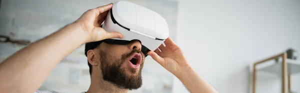 Scioccato uomo barbuto regolazione auricolare vr durante il gioco a casa, banner — Foto stock