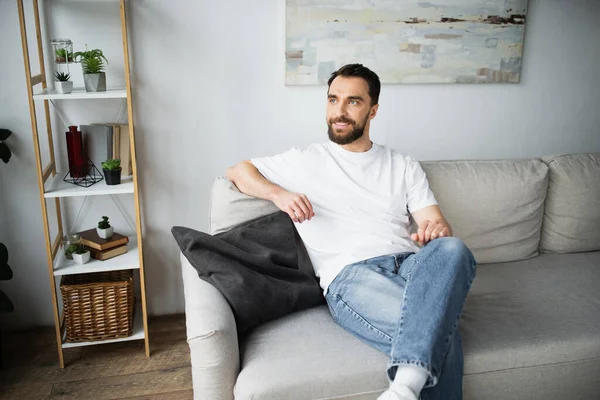 Довольный и бородатый мужчина в джинсах сидит на диване в современной гостиной — стоковое фото