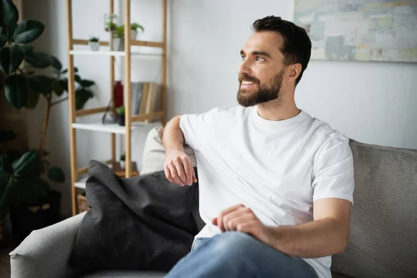 Pleased and bearded man in jeans sitting on couch in modern living room — Stock Photo