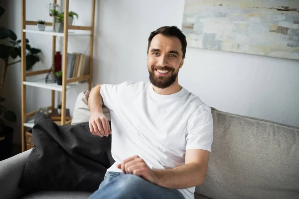 Щасливий і бородатий чоловік у білій футболці, що сидить на дивані в сучасній вітальні — стокове фото