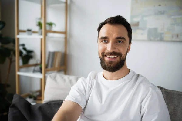 Hombre alegre y barbudo en camiseta blanca sentado en el sofá en la sala de estar moderna - foto de stock