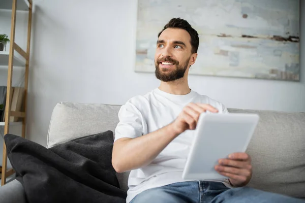Щасливий чоловік у білій футболці сидить на дивані і використовує цифровий планшет у сучасній вітальні — стокове фото