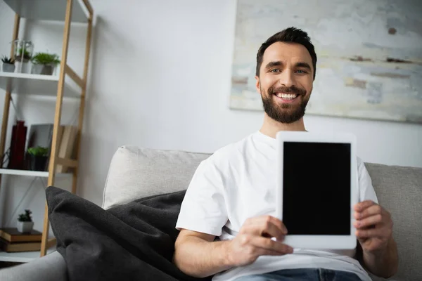 Glücklicher und bärtiger Mann im weißen T-Shirt, der auf der Couch sitzt und ein digitales Tablet mit leerem Bildschirm in der Hand hält — Stockfoto