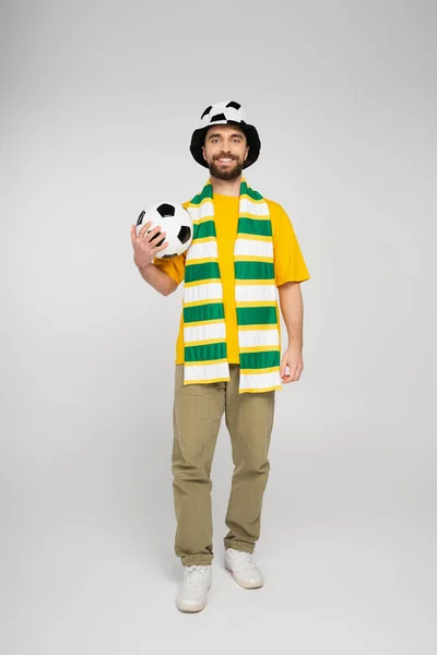 Полный рост счастливого босоногого болельщика в шляпе, держащего футбол на сером — стоковое фото