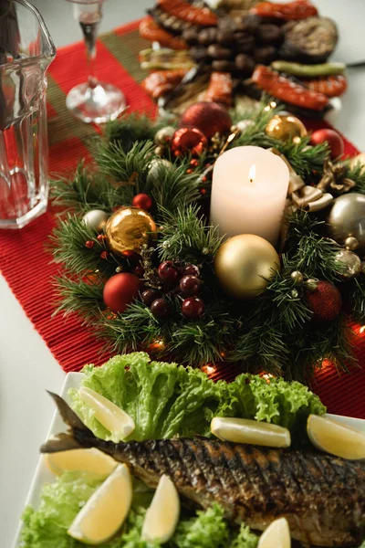 Couronne de Noël avec des boules brillantes et une bougie allumée près de poissons grillés avec du citron tranché servi pour le dîner festif — Photo de stock