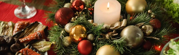 Рождественский венок с горящей свечой и блестящими безделушками возле жареных овощей на столе с праздничным ужином, баннером — стоковое фото