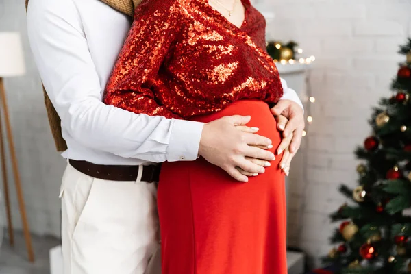 Vista parcial del hombre abrazando la barriga de la mujer embarazada en ropa elegante durante la celebración de Navidad - foto de stock