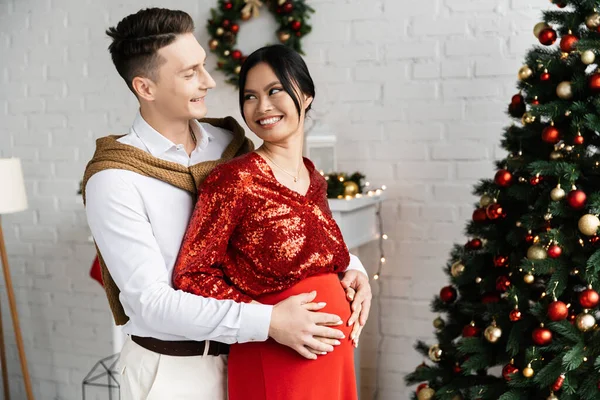 Embarazada asiático mujer sonriendo a feliz marido abrazando su vientre cerca decorado árbol de Navidad - foto de stock